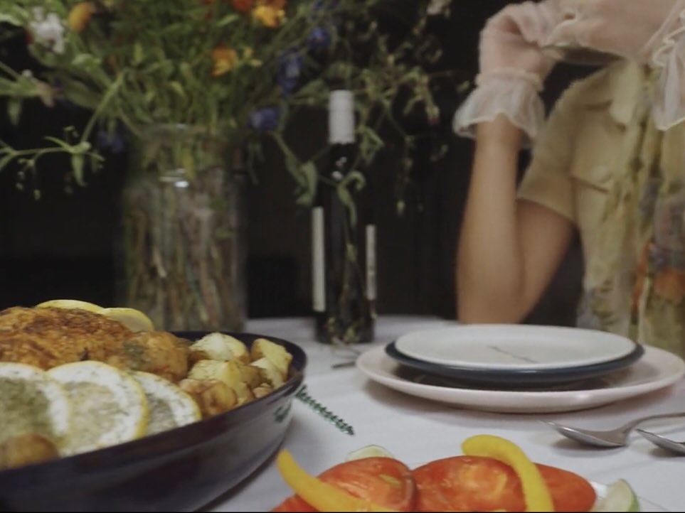 Film still: candlelit table detail — dried flowers, bread and a gloved hand reaching in