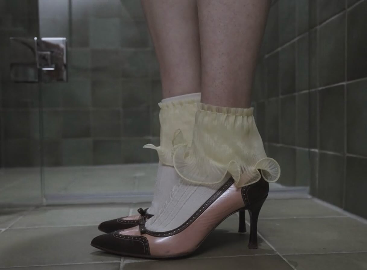 Film still: close-up of frilled socks and two-tone kitten heels on tiled floor
