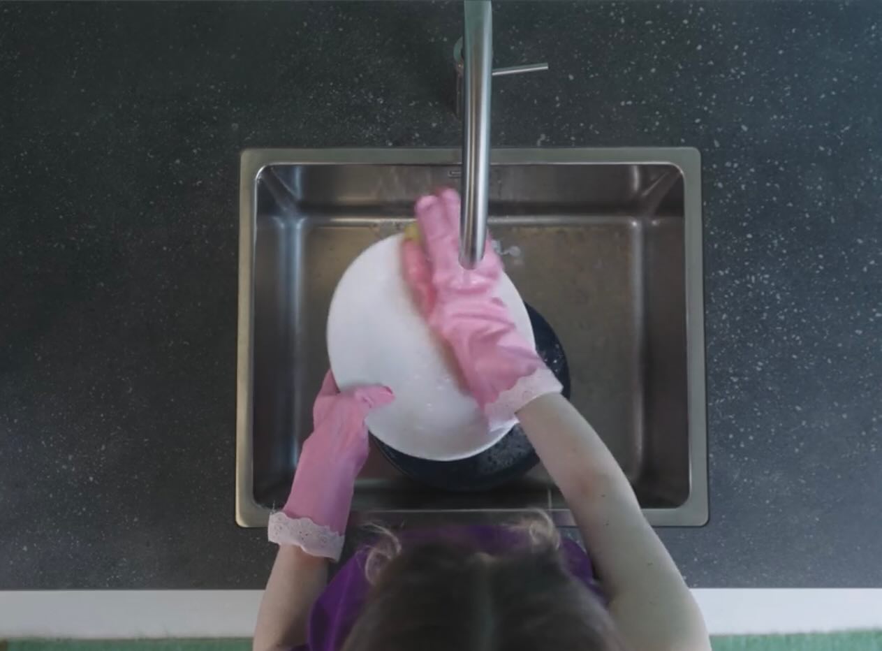 Film still: pink rubber gloves washing a plate under the tap, shot from above