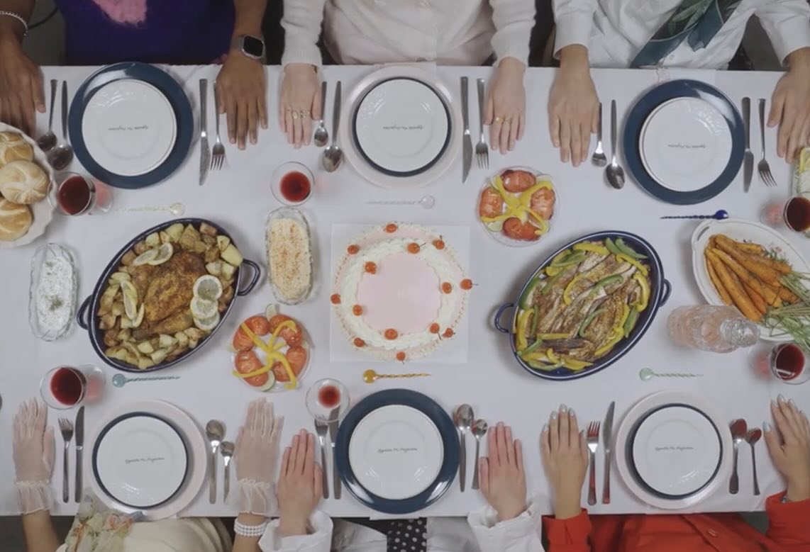 Film still: the dining table laid out from above, gloved hands resting beside each plate